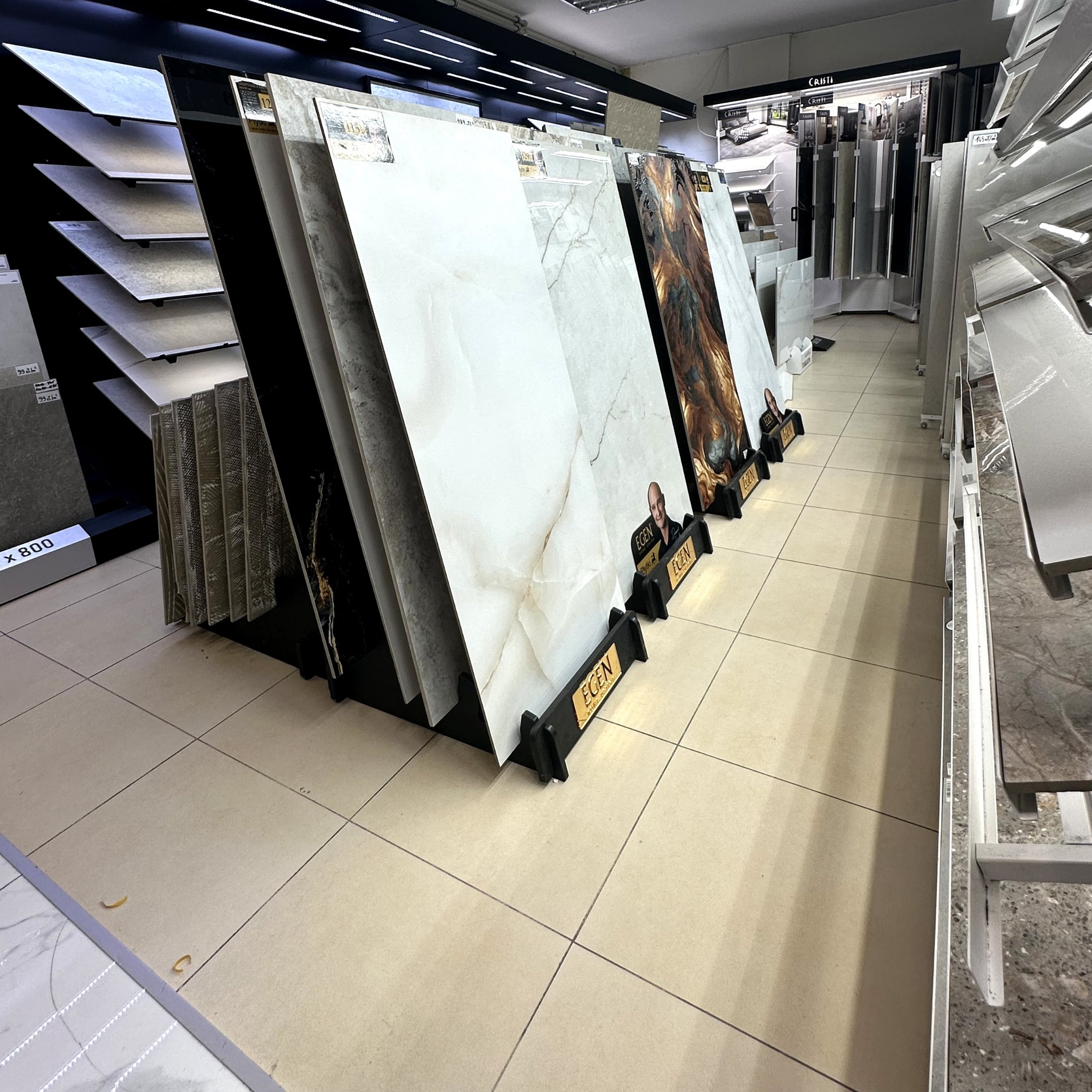 Ekspozycja płytek ceramicznych w sklepie. Widoczne różne wzory i kolory płytek, ustawione pionowo na stojakach z logo 'EGEN'. Oświetlenie sufitowe.