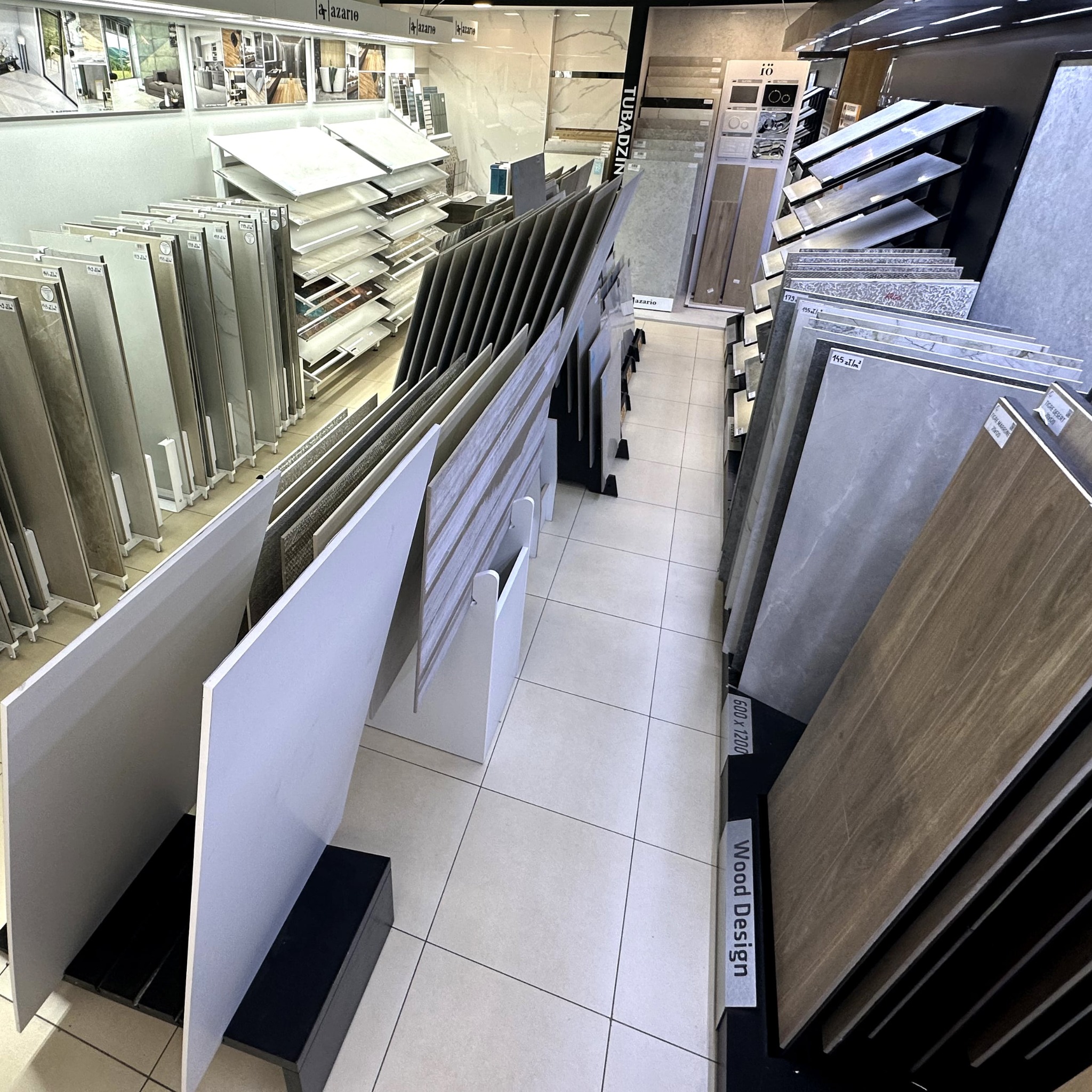 Ekspozycja płytek ceramicznych różnych wzorów w salonie sprzedaży. Widoczne kolekcje Tubądzin, Azario i inne. Płytki ułożone w stojakach.