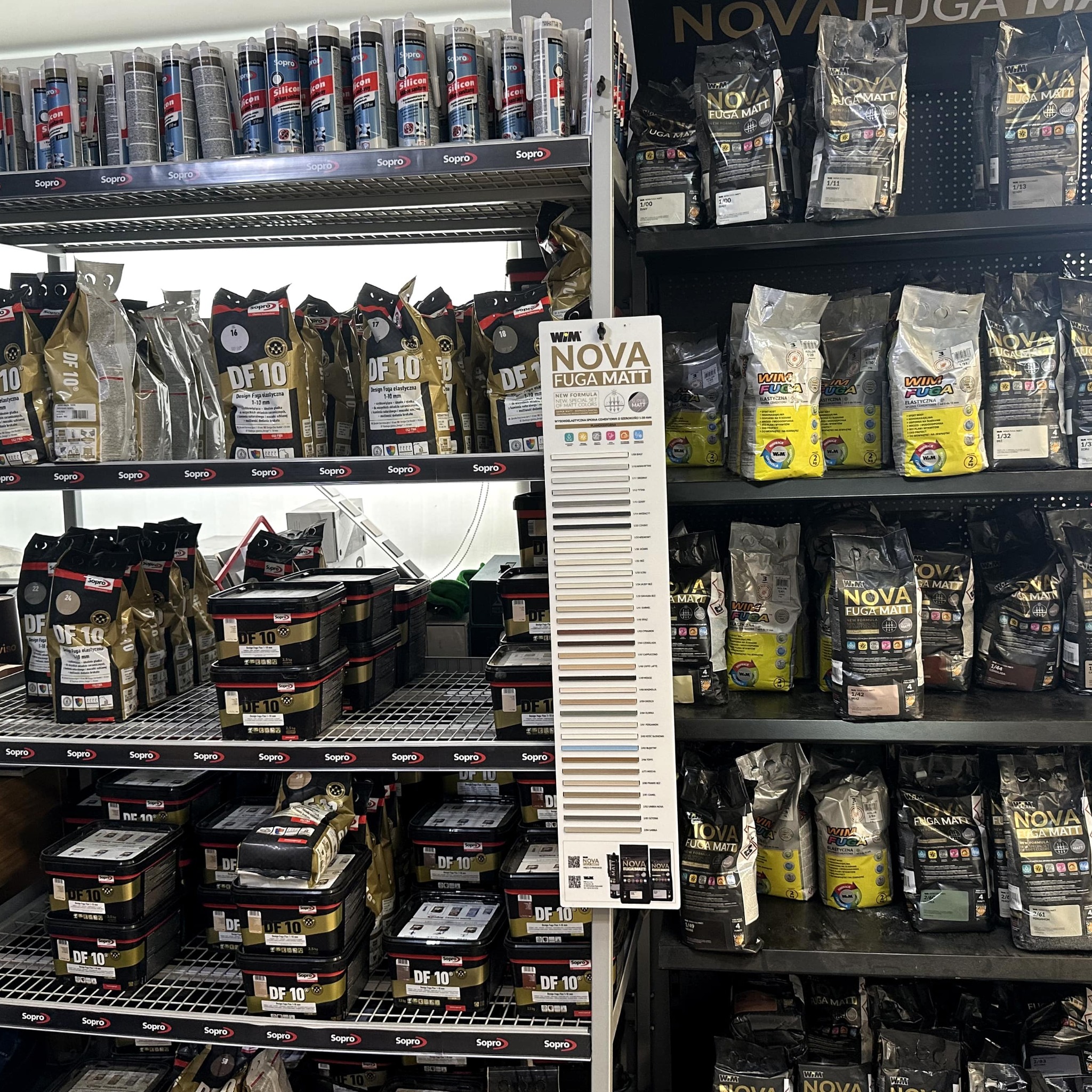 Półki sklepowe z materiałami budowlanymi: opakowania fug Sopro DF 10 i Nova Fuga Matt, silikony. W tle okno, oświetlenie dzienne. Prezentacja kolorów fug na ekspozytorze.