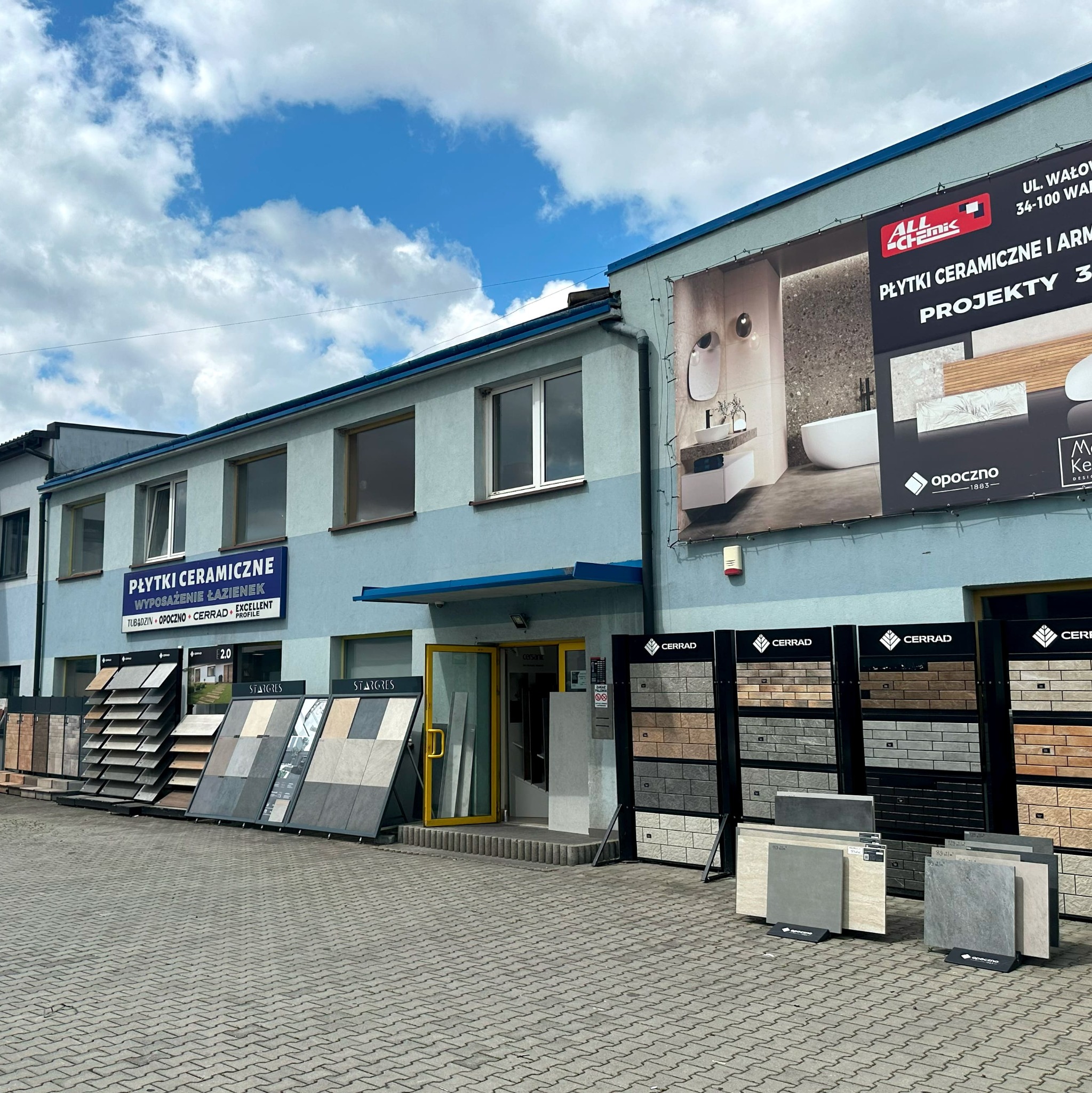 Salon z ekspozycją płytek ceramicznych marek Cerrad i Opoczno. Na budynku baner reklamowy z aranżacją łazienki. Błękitne niebo z chmurami w tle.