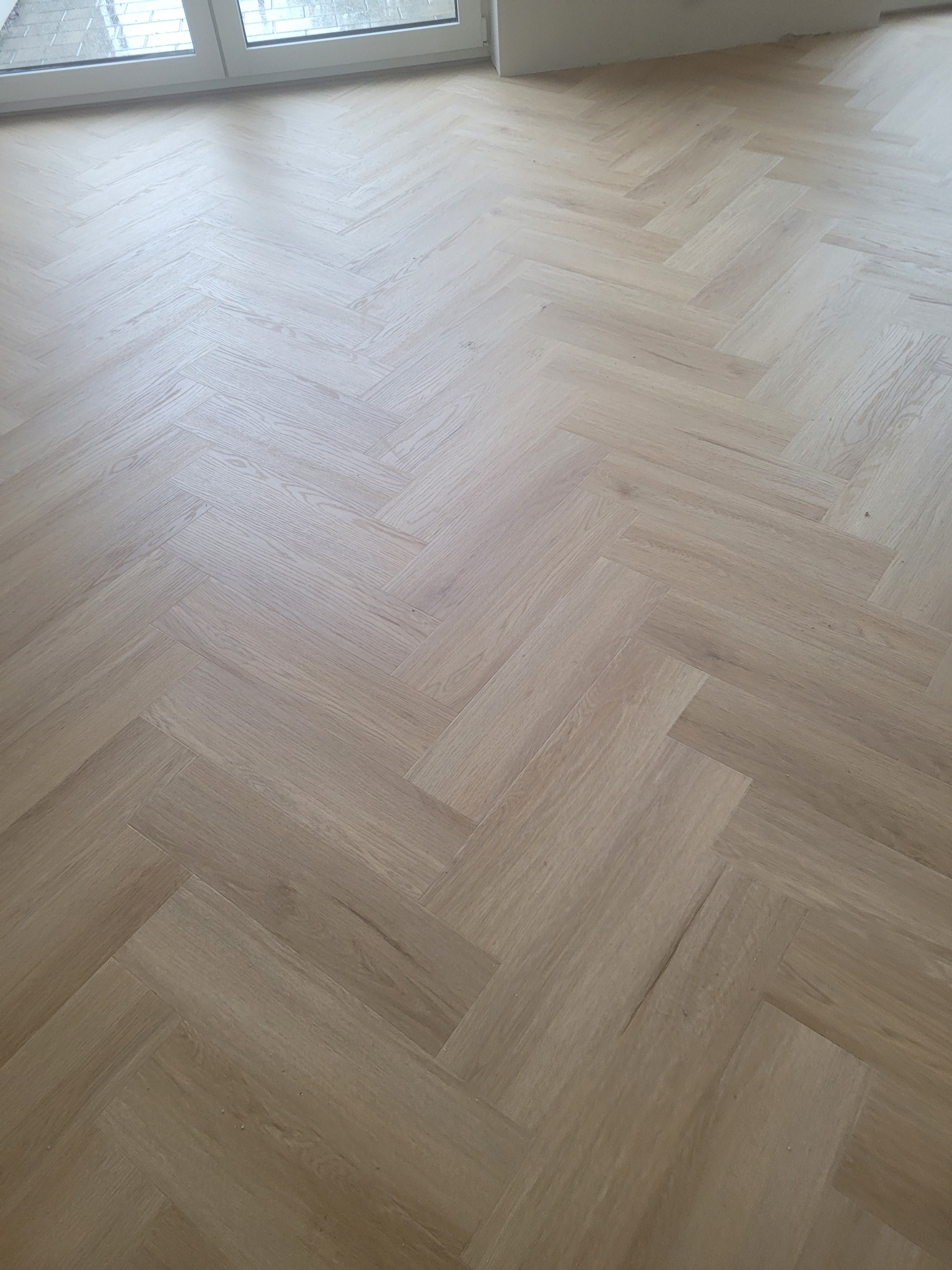 Jasne panele ułożone w jodełkę francuską, widok z góry, blisko okna. Naturalne światło podkreśla strukturę drewna, tworząc ciepły i nowoczesny wygląd podłogi.