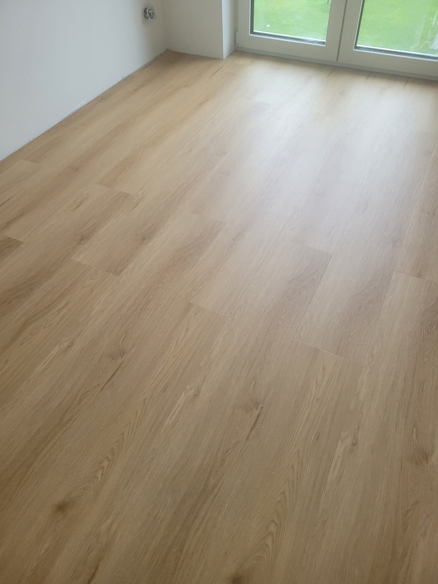 Jasne panele podłogowe ułożone w nowym pomieszczeniu z białymi ścianami i oknem, przez które widać zieleń. Widok z góry, bliski kadr.