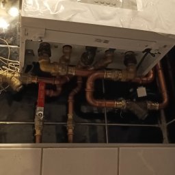 Pawel Pisula - Świeżo zamontowany, biały piec gazowy z miedzianymi rurami na tle czarno-białych płytek. Widoczny zawór Kros i gniazdko elektryczne. Ujęcie z dołu.