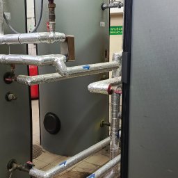 Pawel Pisula - Szczegółowe ujęcie instalacji grzewczej z izolowanymi rurami, manometrami i zaworami w kotłowni. Widoczne elementy systemu grzewczego i rury z oznaczeniami kierunku przepływu.