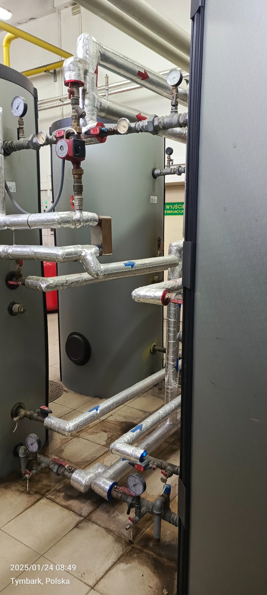 Szczegółowe ujęcie instalacji grzewczej z izolowanymi rurami, manometrami i zaworami w kotłowni. Widoczne elementy systemu grzewczego i rury z oznaczeniami kierunku przepływu.