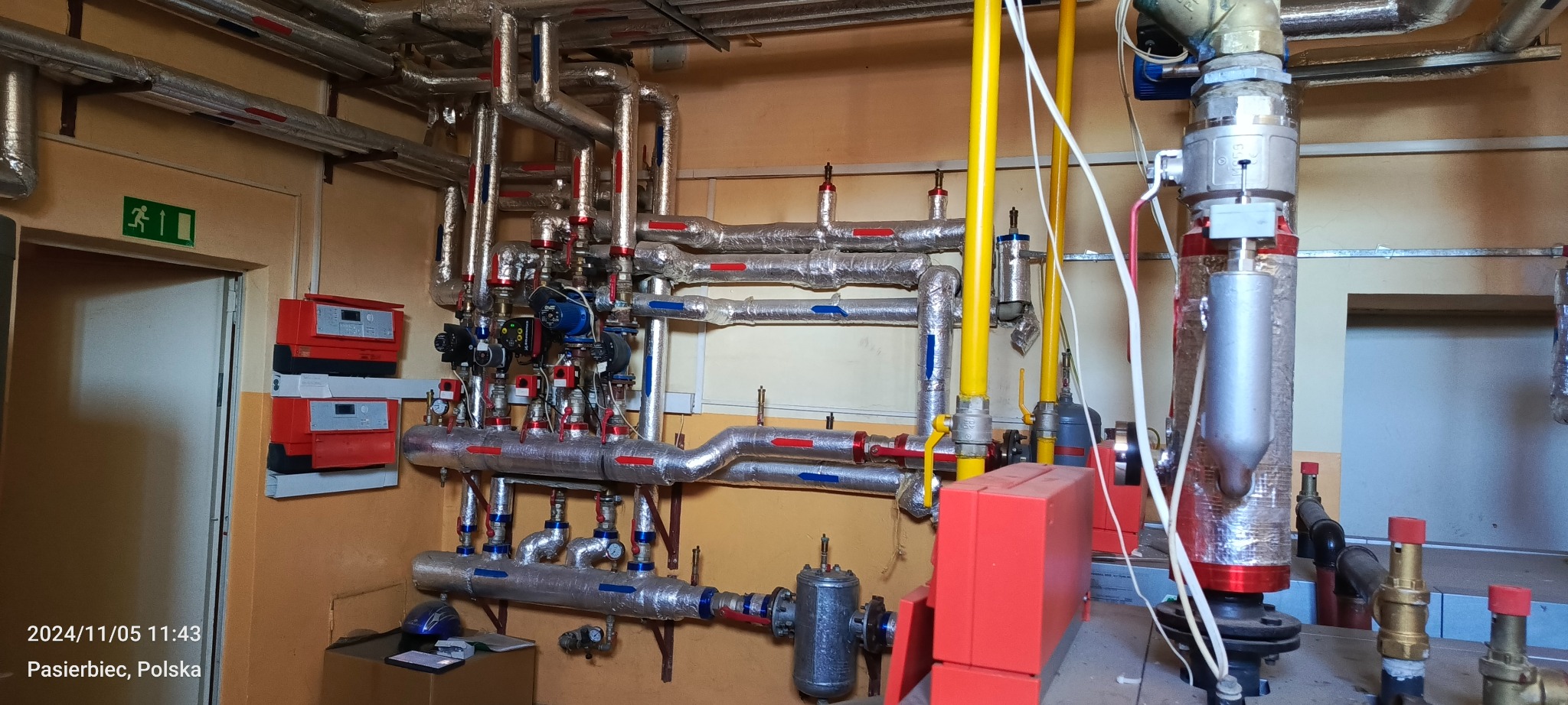 Złożona instalacja grzewcza z rurami w srebrnej izolacji, czerwonymi i niebieskimi oznaczeniami. Widoczne pompy, zawory i sterowniki. Instalacja w pomieszczeniu o żółtych ścianach.