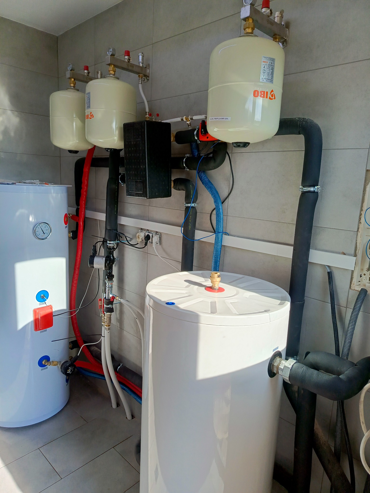 Białe zbiorniki ciśnieniowe IBO z rurami i zaworami na tle szarej ściany z płytek. Instalacja w pomieszczeniu technicznym, widoczne elementy instalacji grzewczej.