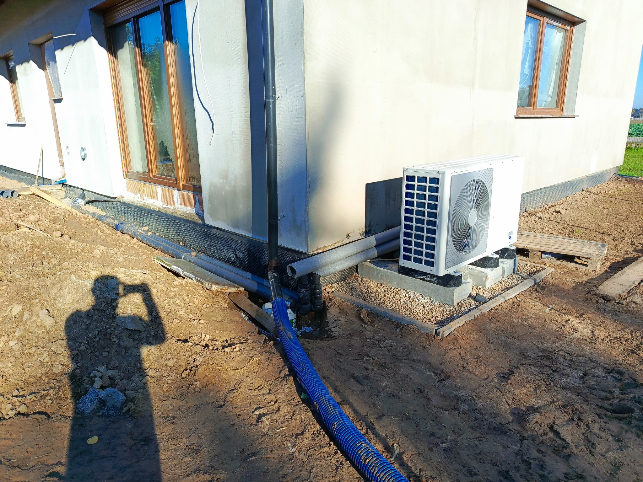 Biała pompa ciepła na zewnątrz budynku, z widocznymi rurami i shadow osoby robiącej zdjęcie. Instalacja w trakcie prac ziemnych.