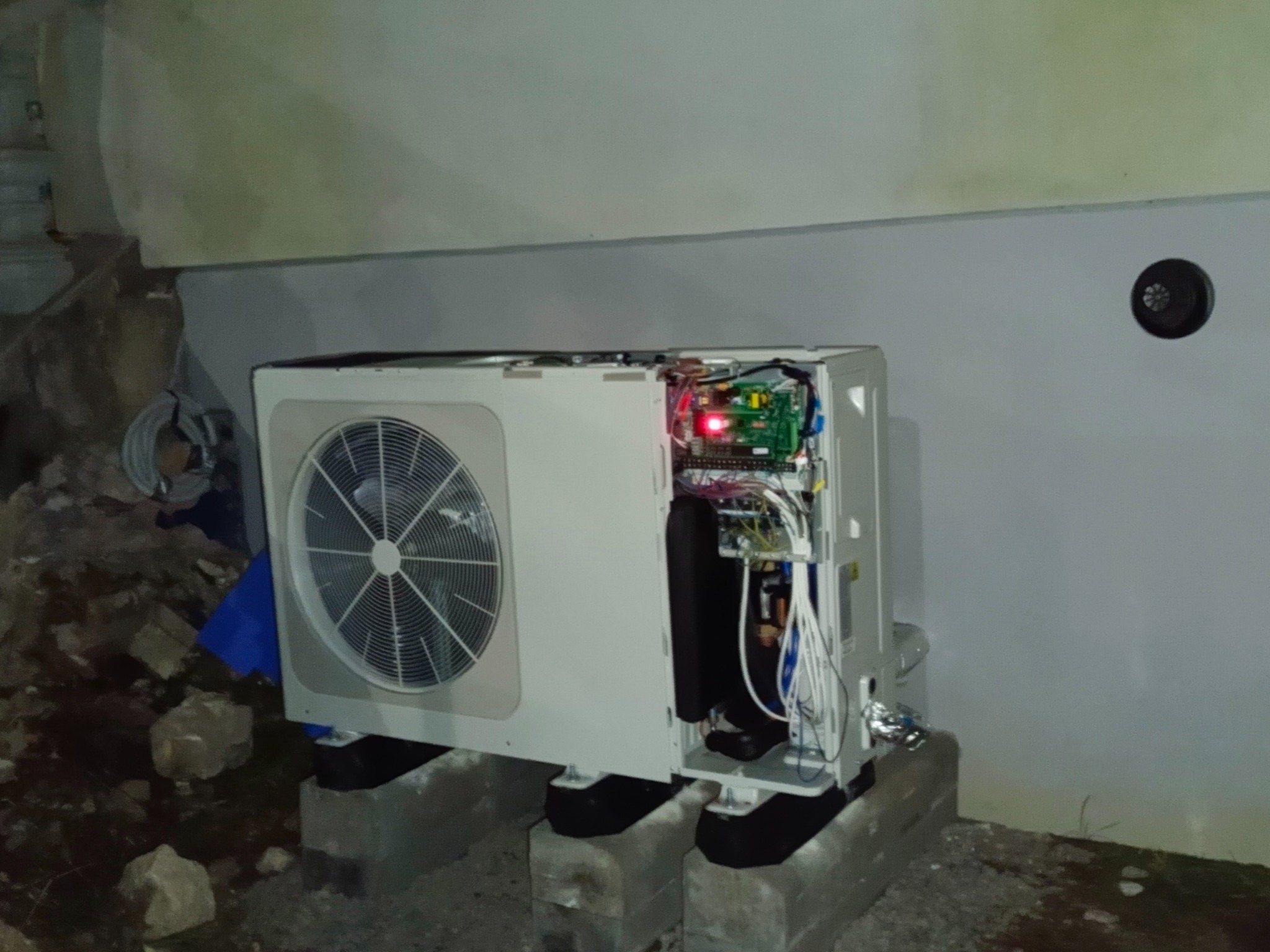 Otwarta obudowa pompy ciepła na betonowych blokach, widoczne wewnętrzne komponenty i okablowanie. Tło stanowi szara ściana i gruz.