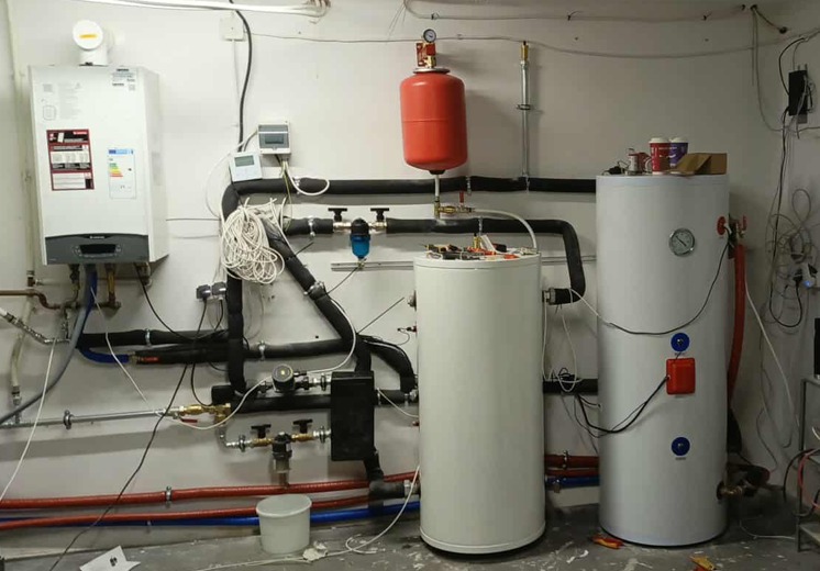 Instalacja grzewcza w kotłowni: piec gazowy, dwa zbiorniki, rury w izolacji, manometry, zawory. Widok na ścianę z elementami instalacji.