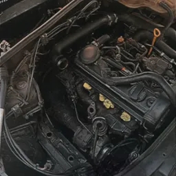 Otwarta maska samochodu z widocznym silnikiem. Elementy silnika pokryte żółtymi naklejkami. Widoczne przewody, zbiorniki i inne komponenty.