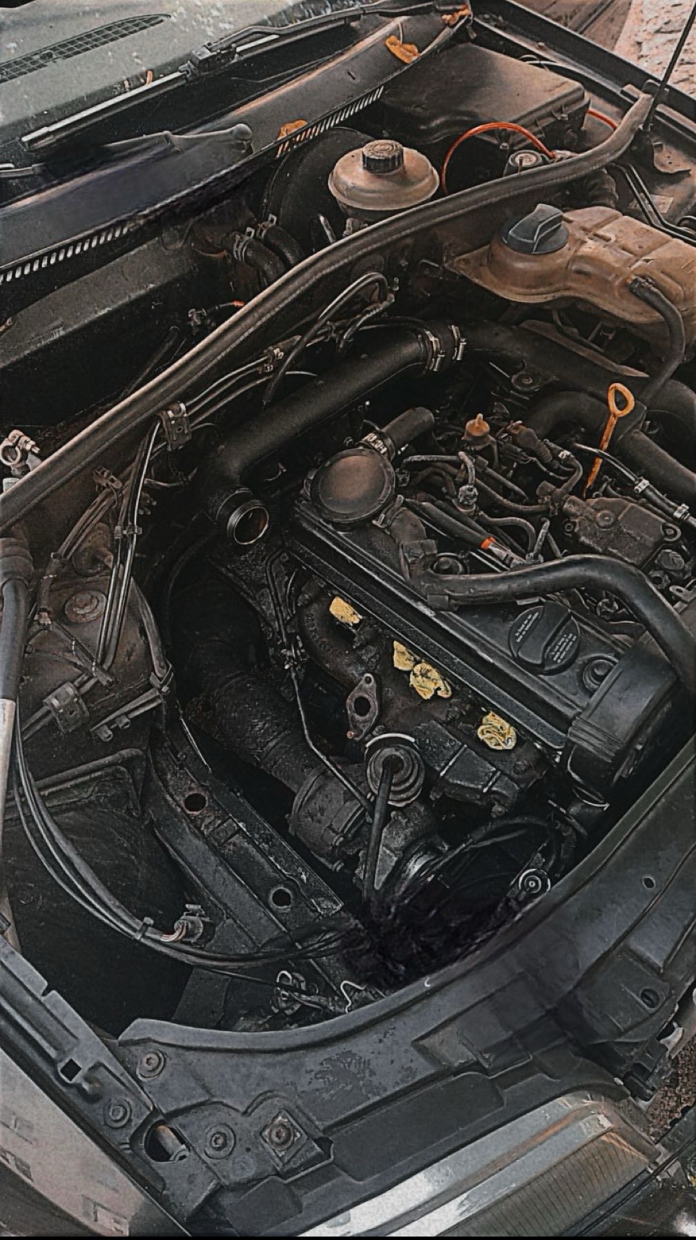 Otwarta maska samochodu z widocznym silnikiem. Elementy silnika pokryte żółtymi naklejkami. Widoczne przewody, zbiorniki i inne komponenty.