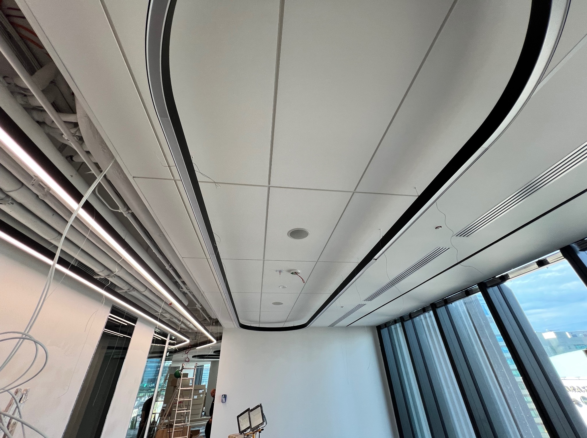 Kasetonowy sufit podwieszany z oświetleniem LED, widok z dołu. Elementy instalacji widoczne nad sufitem. Duże okna z widokiem na miasto.