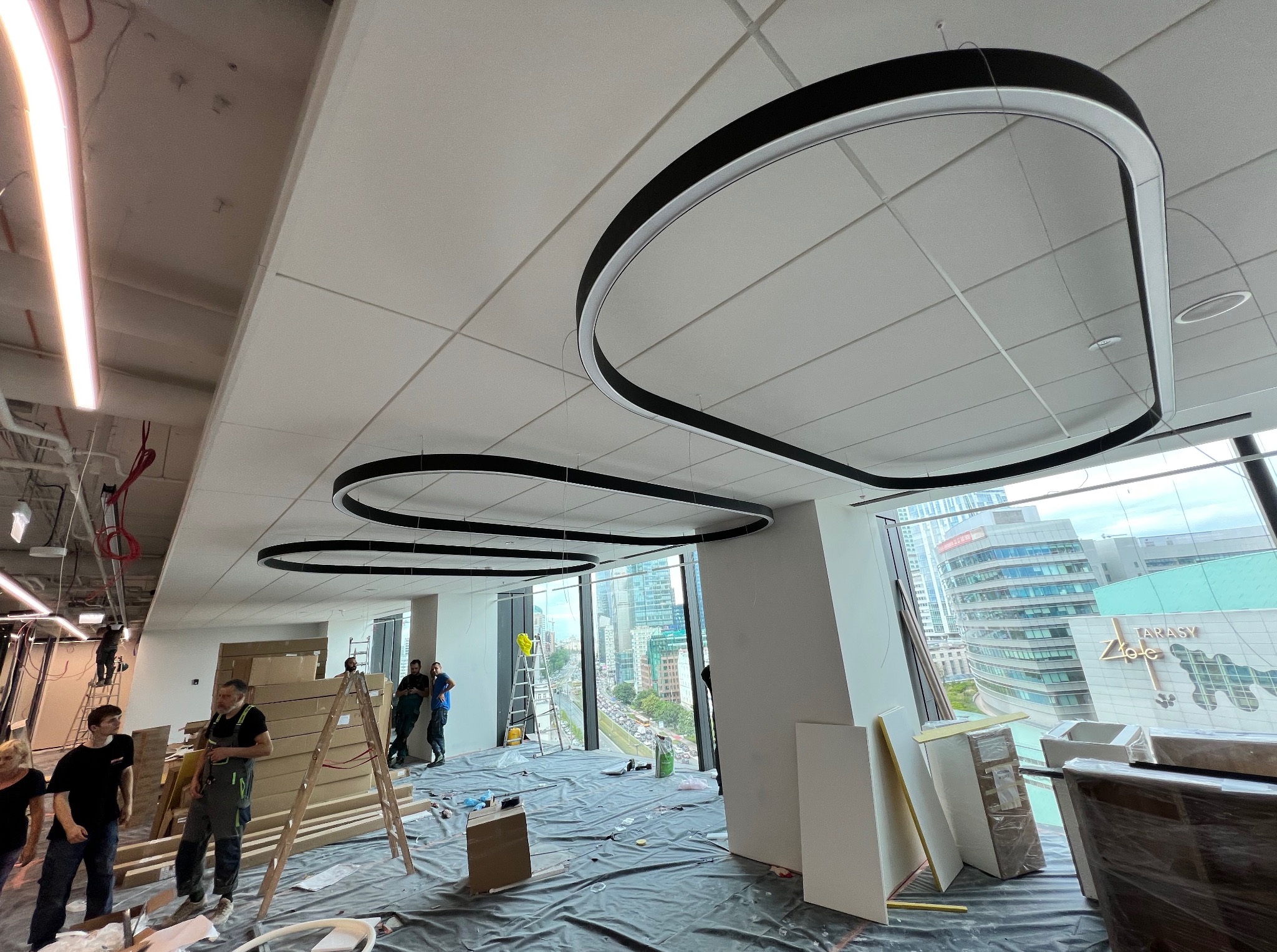 Montaż sufitu podwieszanego z oświetleniem w biurze w Warszawie. Widoczne płyty i ekipa montażowa. Duże okna z widokiem na miasto.