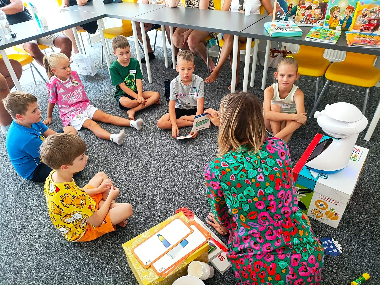 Grupa dzieci siedzi na podłodze w kręgu, słuchając nauczycielki podczas lekcji języka angielskiego z użyciem kart obrazkowych i edukacyjnego robota.