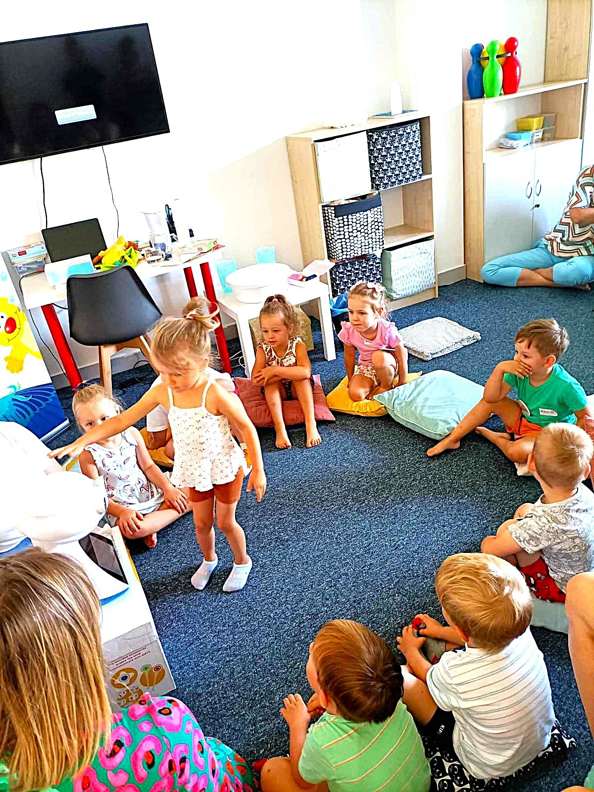 Grupa dzieci siedzi w kręgu na dywanie podczas zajęć w jasnym pomieszczeniu. Dziewczynka w białej koszulce gestykuluje, prowadząc zabawę. W tle widoczne półki z zabawkami.