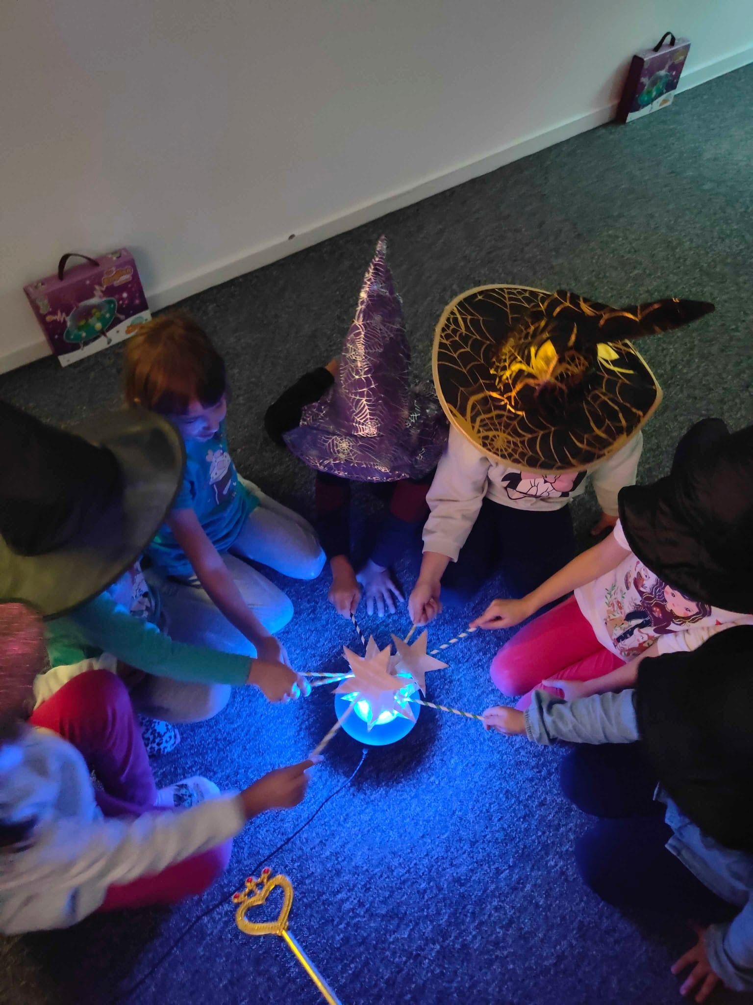 Dzieci w czapkach czarownic siedzą w kręgu, trzymając różdżki nad świecącą kulą. Zabawa edukacyjna w szkole językowej dla dzieci.