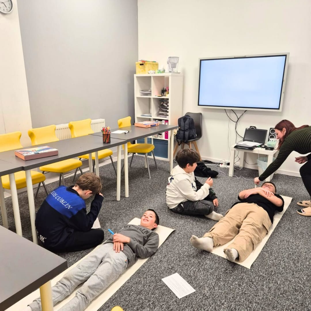 Lekcja relaksacji dla dzieci w szkole językowej: chłopcy leżą na matach, nauczycielka pokazuje ćwiczenie, w tle stoliki i krzesła.