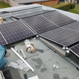 LUXGRID SYSTEMS SPÓŁKA Z OGRANICZONĄ ODPOWIEDZIALNOŚCIĄ - Montaż paneli fotowoltaicznych na dachu budynku. Widoczne panele, elementy montażowe i narzędzia. Tło stanowią budynki mieszkalne i drzewa.