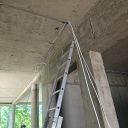 LUXGRID SYSTEMS SPÓŁKA Z OGRANICZONĄ ODPOWIEDZIALNOŚCIĄ - Wnętrze w surowym stanie z przewodami elektrycznymi przymocowanymi do sufitu i drabina oparta o filar. Widoczny fragment zieleni za oknem i sylwetka osoby.