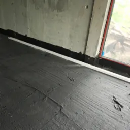 Hydroizolacja podłogi w surowym wnętrzu z oknem. Widoczna czarna masa hydroizolacyjna i biała listwa przypodłogowa. Surowy beton na ścianach.