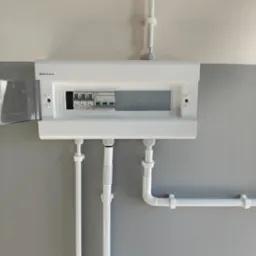 Biała skrzynka elektryczna z otwartymi drzwiczkami, widoczne zabezpieczenia. Instalacja na szarej ścianie, białe rury prowadzą do skrzynki. Widoczne logo Elektro-Plast.