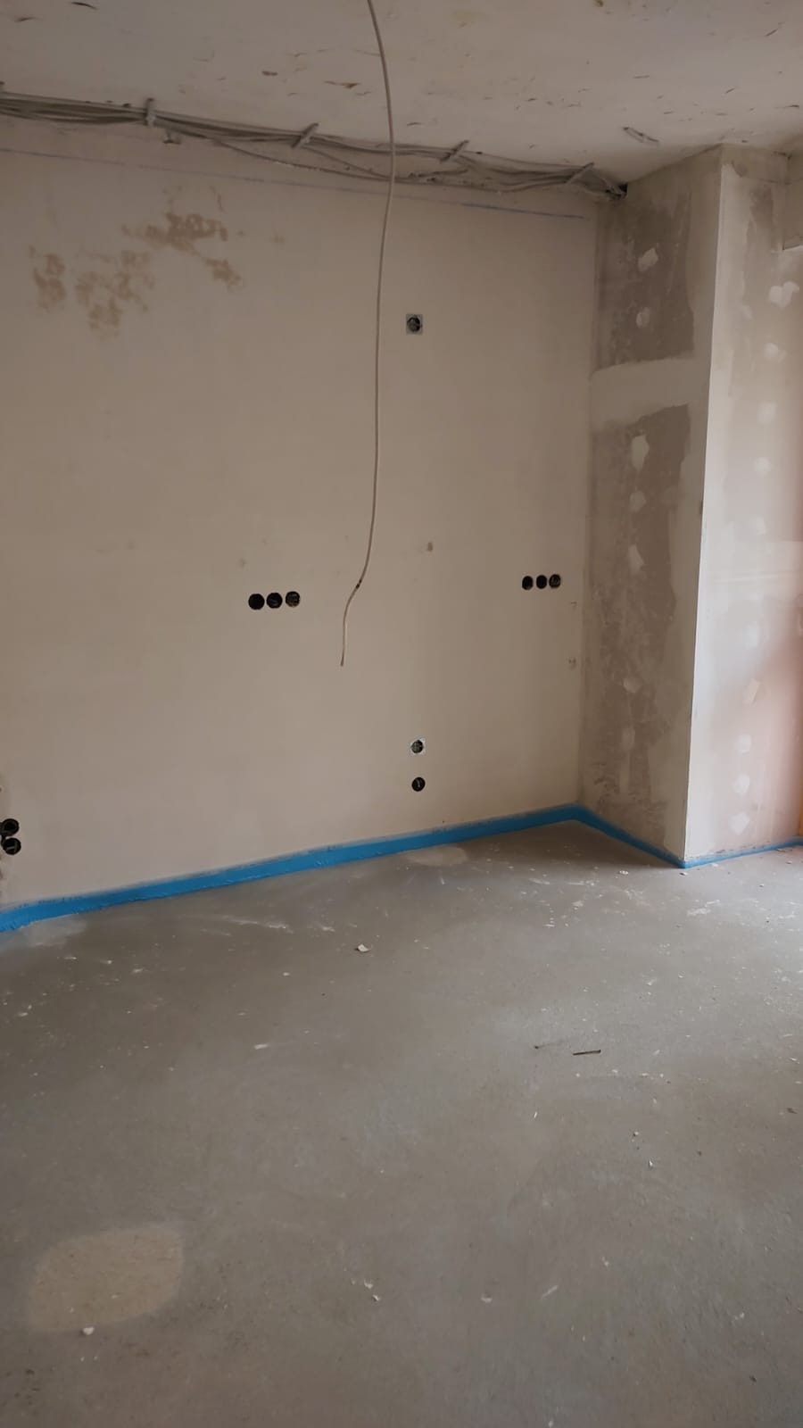 Surowy stan wykończenia wnętrza z widocznymi przewodami elektrycznymi, puszkami instalacyjnymi i zabezpieczeniem podłogi niebieską taśmą malarską.
