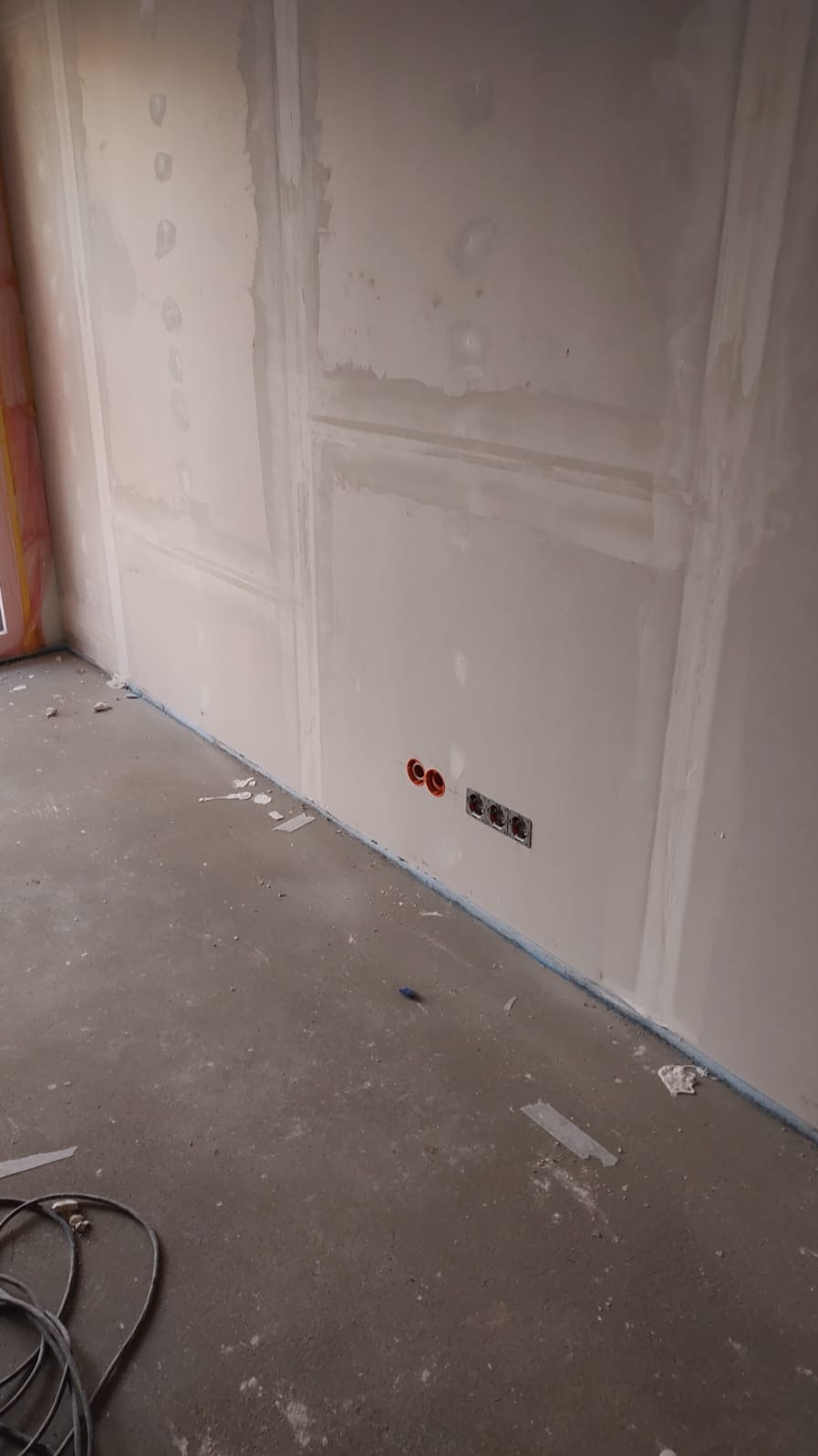 Ściana w trakcie prac wykończeniowych z widocznymi gniazdkami elektrycznymi i puszkami, przygotowana do dalszych prac. Na podłodze leżą kable.
