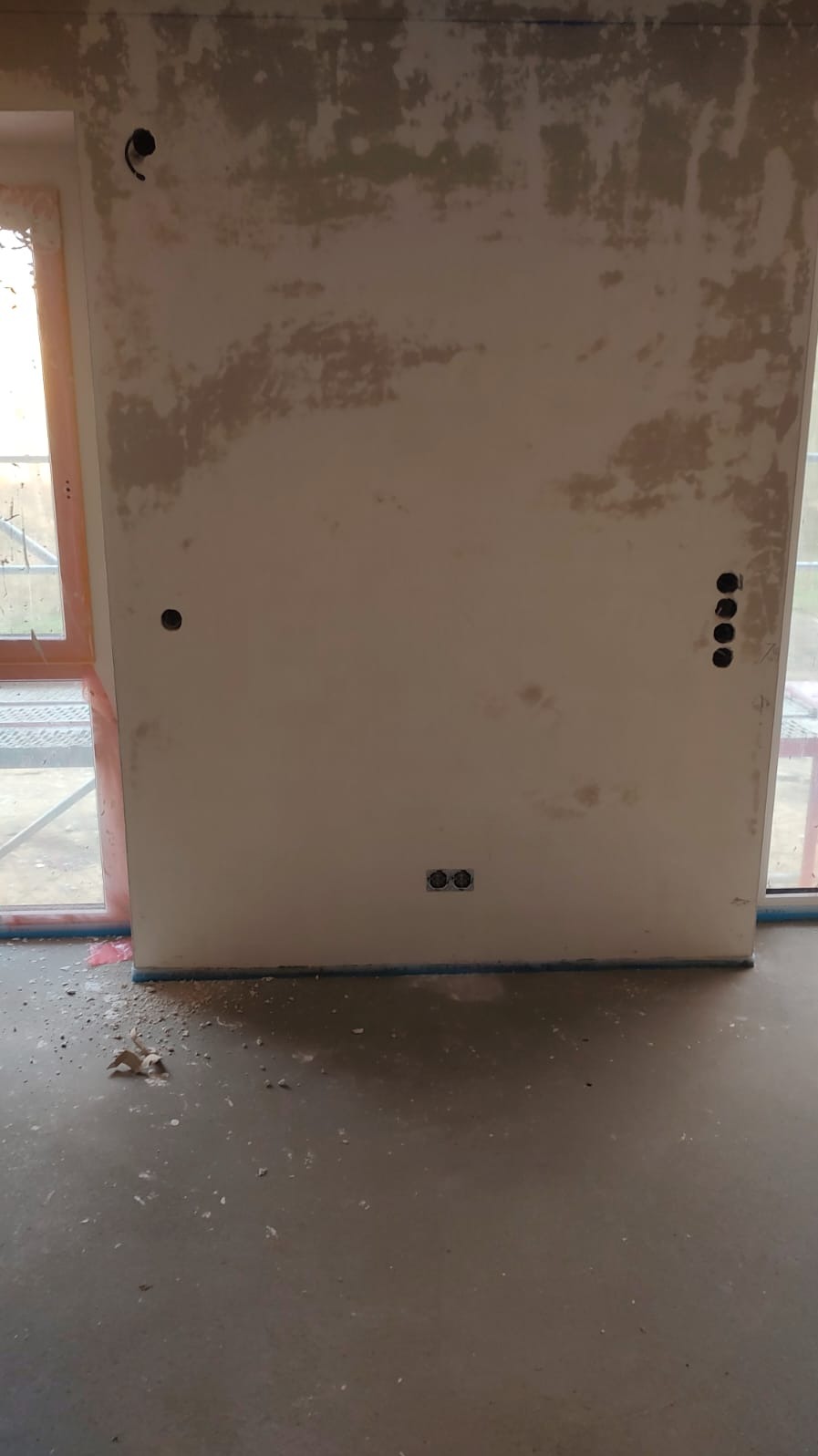 Ściana w trakcie remontu z widocznymi puszkami elektrycznymi, przygotowana do dalszych prac wykończeniowych. Widoczne ślady po starych powłokach i nowe otwory.