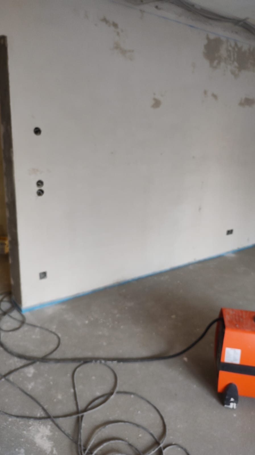 Ściana w trakcie prac wykończeniowych z widocznymi punktami elektrycznymi i kablem na podłodze, pomarańczowe urządzenie w prawym dolnym rogu.