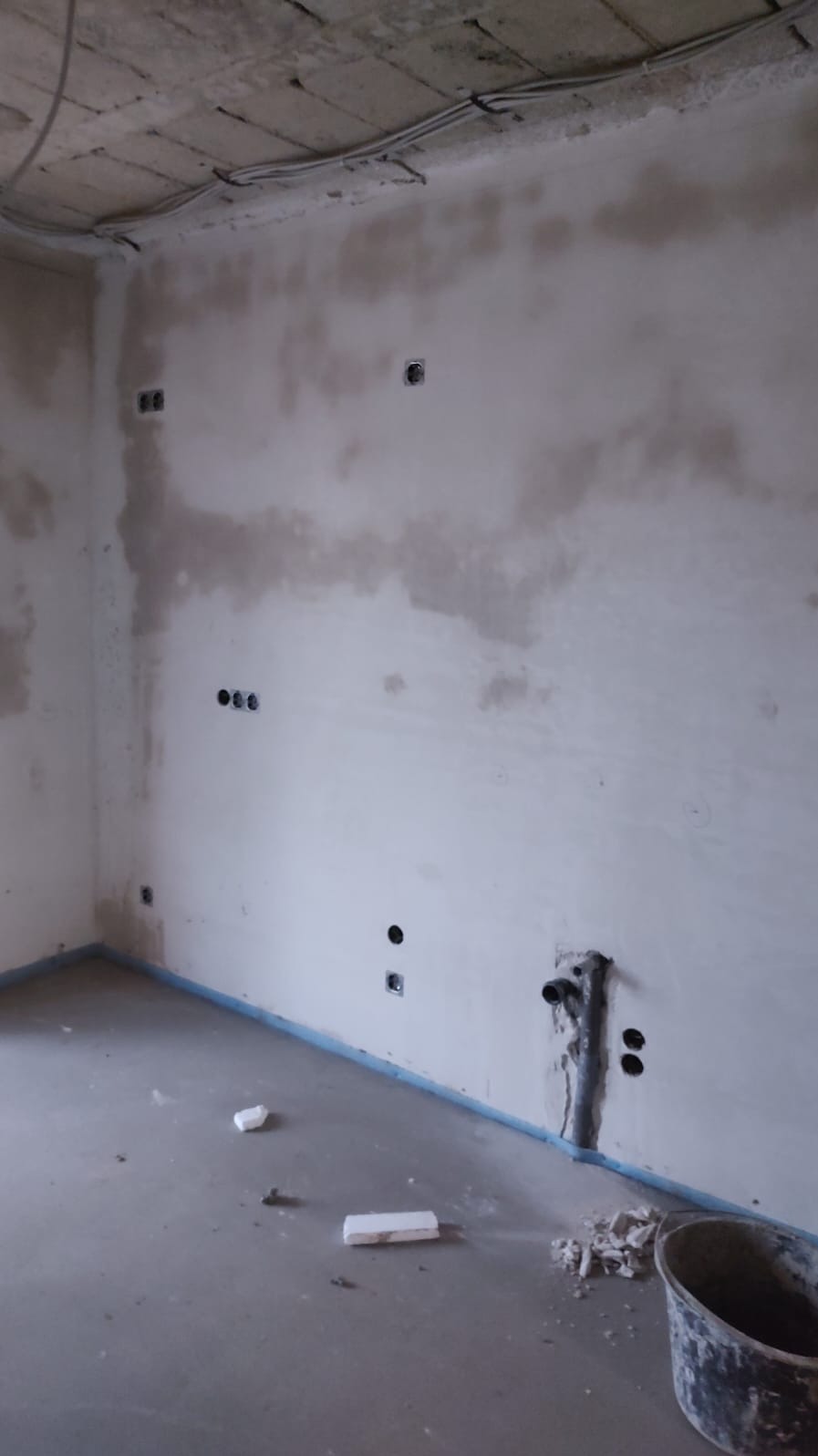 Surowy pokój z instalacją elektryczną i rurami w ścianie, przygotowany do wykończenia. Widoczne kable pod sufitem i otwory na gniazdka.