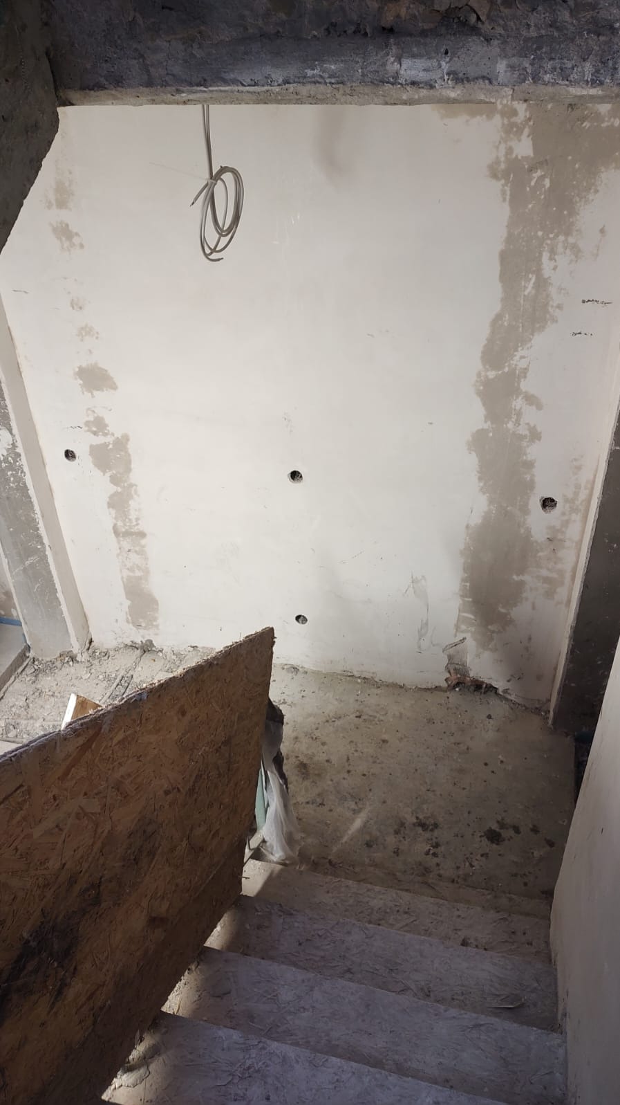 Wnętrze w trakcie remontu: surowe ściany z widocznymi śladami po instalacjach, wystające przewody elektryczne, schody w stanie surowym, zabezpieczone płytą OSB.