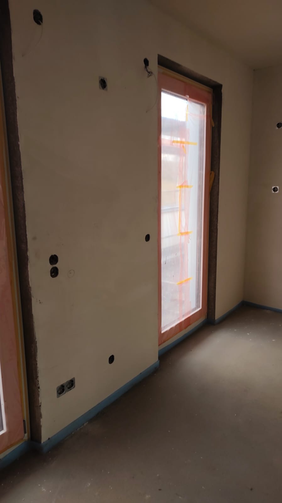Wnętrze w trakcie prac wykończeniowych: ściany z widocznymi puszkami instalacji elektrycznej, okno zabezpieczone folią malarską, surowa posadzka.