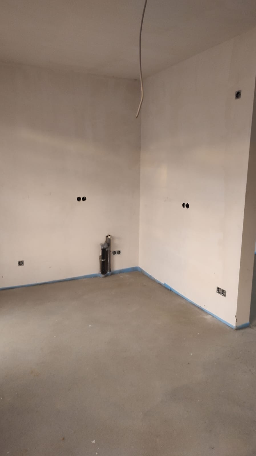 Surowe wnętrze przed wykończeniem: ściany z instalacją elektryczną i rurami, zabezpieczone podłogi taśmą malarską. Widoczne punkty elektryczne i wystające kable.