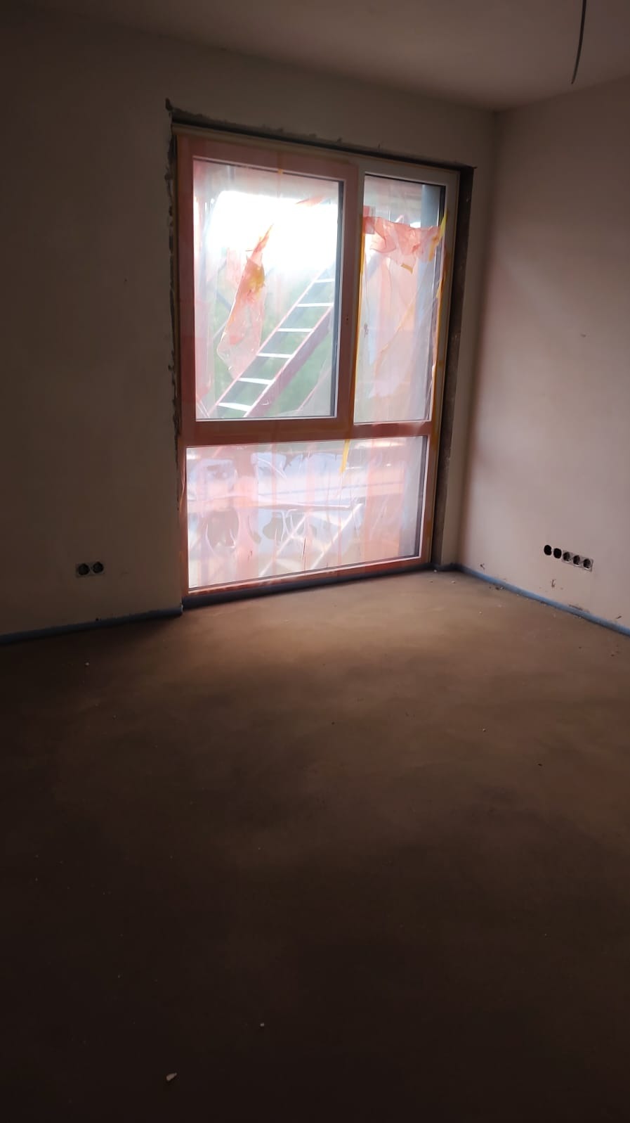 Wnętrze w trakcie prac wykończeniowych: wylewka podłogowa, okno zabezpieczone folią, widoczne puszki elektryczne w ścianie. W tle drabina przez okno.