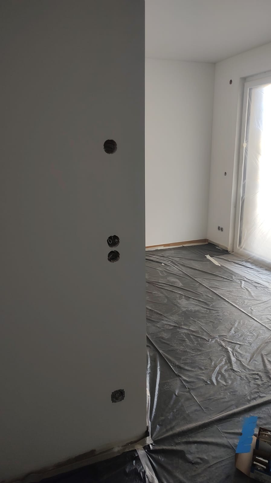 Przygotowanie do montażu gniazdek elektrycznych w nowym mieszkaniu, ściany pomalowane na biało i szaro, podłoga zabezpieczona folią ochronną.
