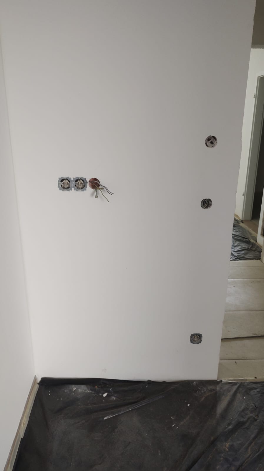 Biała ściana z zamontowanymi puszkami elektrycznymi i wystającymi przewodami. Podłoga zabezpieczona czarną folią malarską. Widoczne gniazdka elektryczne.