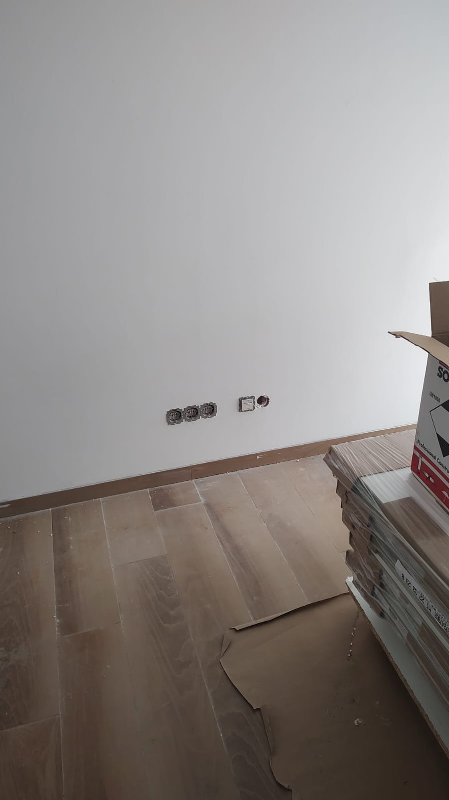 Biała ściana z trzema gniazdkami i jednym włącznikiem bez osprzętu, brązowe panele podłogowe i karton z panelami. Widok z dołu.