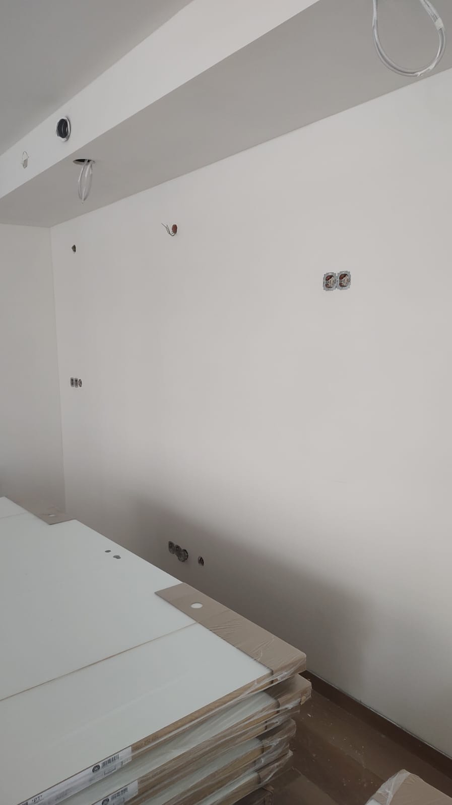 Białe ściany w trakcie remontu, widoczne gniazdka elektryczne, kable wystające z sufitu i stos płyt meblowych z zabezpieczeniami na rogach.