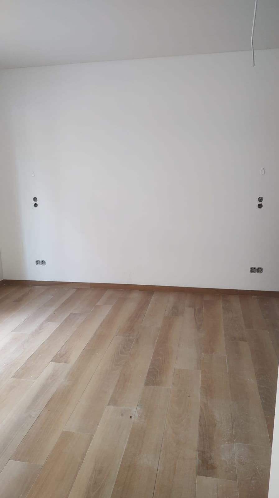 Jasny parkiet w pustym pokoju z białymi ścianami, na których widoczne są puszki elektryczne na różnych wysokościach. Surowy stan przed montażem osprzętu.