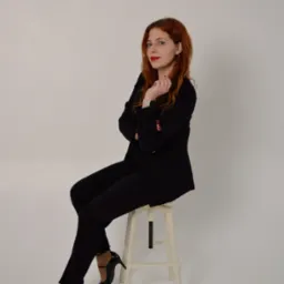 Portret kobiety z rudymi włosami w czarnym garniturze, siedzącej na białym stołku na jasnym tle. Uwagę przyciąga jej pewny wyraz twarzy i elegancki strój.