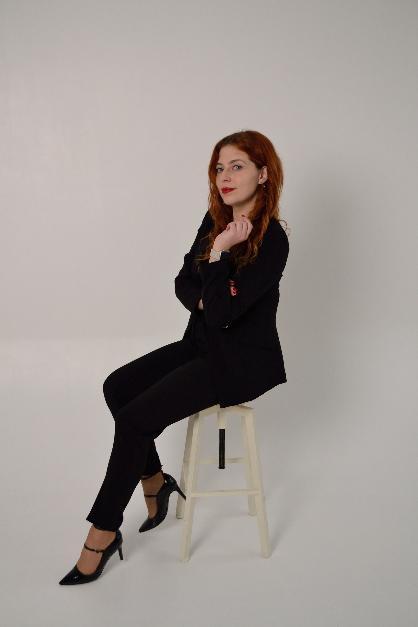 Portret kobiety z rudymi włosami w czarnym garniturze, siedzącej na białym stołku na jasnym tle. Uwagę przyciąga jej pewny wyraz twarzy i elegancki strój.