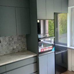 Meble Białek - Szare, matowe szafki kuchenne z mozaiką nad blatem i wbudowanym piekarnikiem, widok z boku, naturalne światło z okna. Minimalistyczny design.