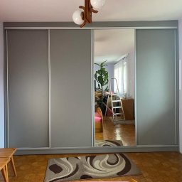 Meble Białek - Szafa wnękowa z przesuwnymi, szarymi drzwiami i lustrem, w którym odbija się pokój z drabiną i roślinami. Na podłodze wzorzysty dywan.