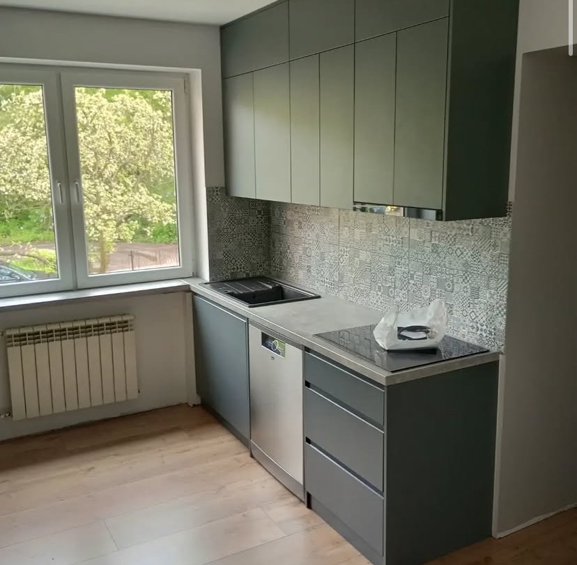 Nowoczesna kuchnia z szarymi szafkami, wzorzystą mozaiką na ścianie i jasnym drewnem na podłodze. Okno z widokiem na zieleń dodaje naturalnego światła.
