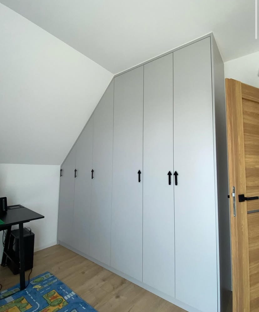 Jasnoszara szafa wnękowa z czarnymi uchwytami, dopasowana do skosu dachu. Widok z boku na drewnianą ościeżnicę drzwi i fragment pokoju.