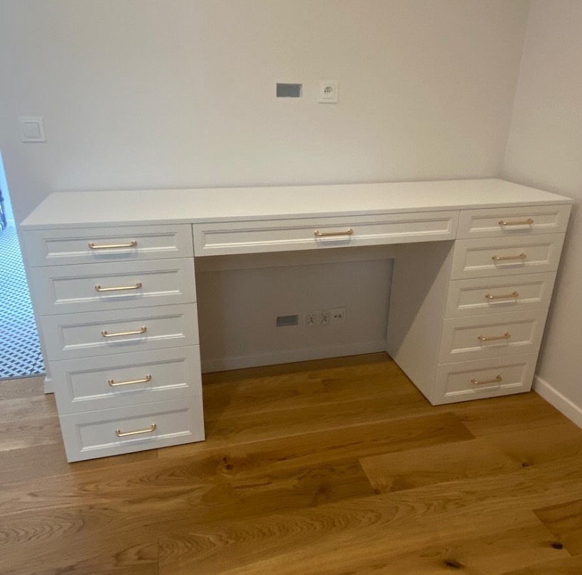Białe biurko na wymiar z szufladami i złotymi uchwytami, ustawione na drewnianej podłodze. Widoczne gniazdka elektryczne pod blatem.