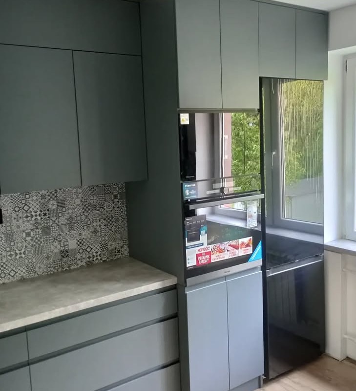 Szare, matowe szafki kuchenne z mozaiką nad blatem i wbudowanym piekarnikiem, widok z boku, naturalne światło z okna. Minimalistyczny design.
