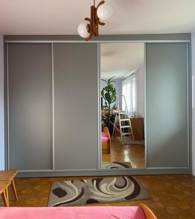 Szafa wnękowa z przesuwnymi, szarymi drzwiami i lustrem, w którym odbija się pokój z drabiną i roślinami. Na podłodze wzorzysty dywan.