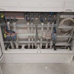 Rozdzielnica elektryczna w szarej obudowie z widocznymi kablami, listwami zaciskowymi i elementami montażowymi. Widok z góry na otwartą skrzynkę.